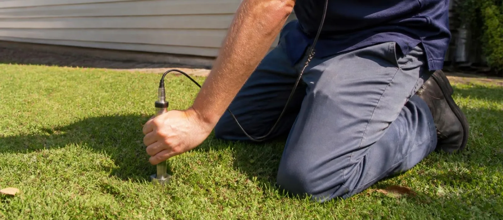 Technician kneeling on a lawn pressing a listening probe into the ground to find a buried leak