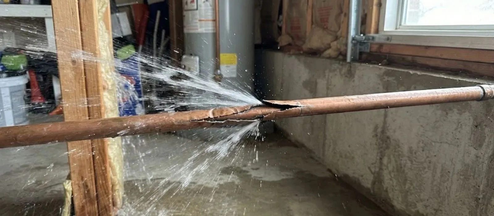 Burst copper pipe spraying water along a basement wall beside a water heater