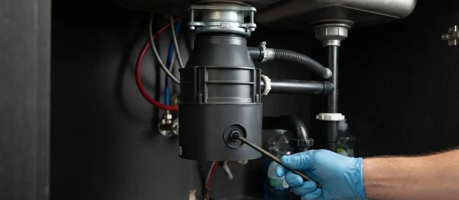 Gloved hand using a wrench on a garbage disposal mounted under a kitchen sink